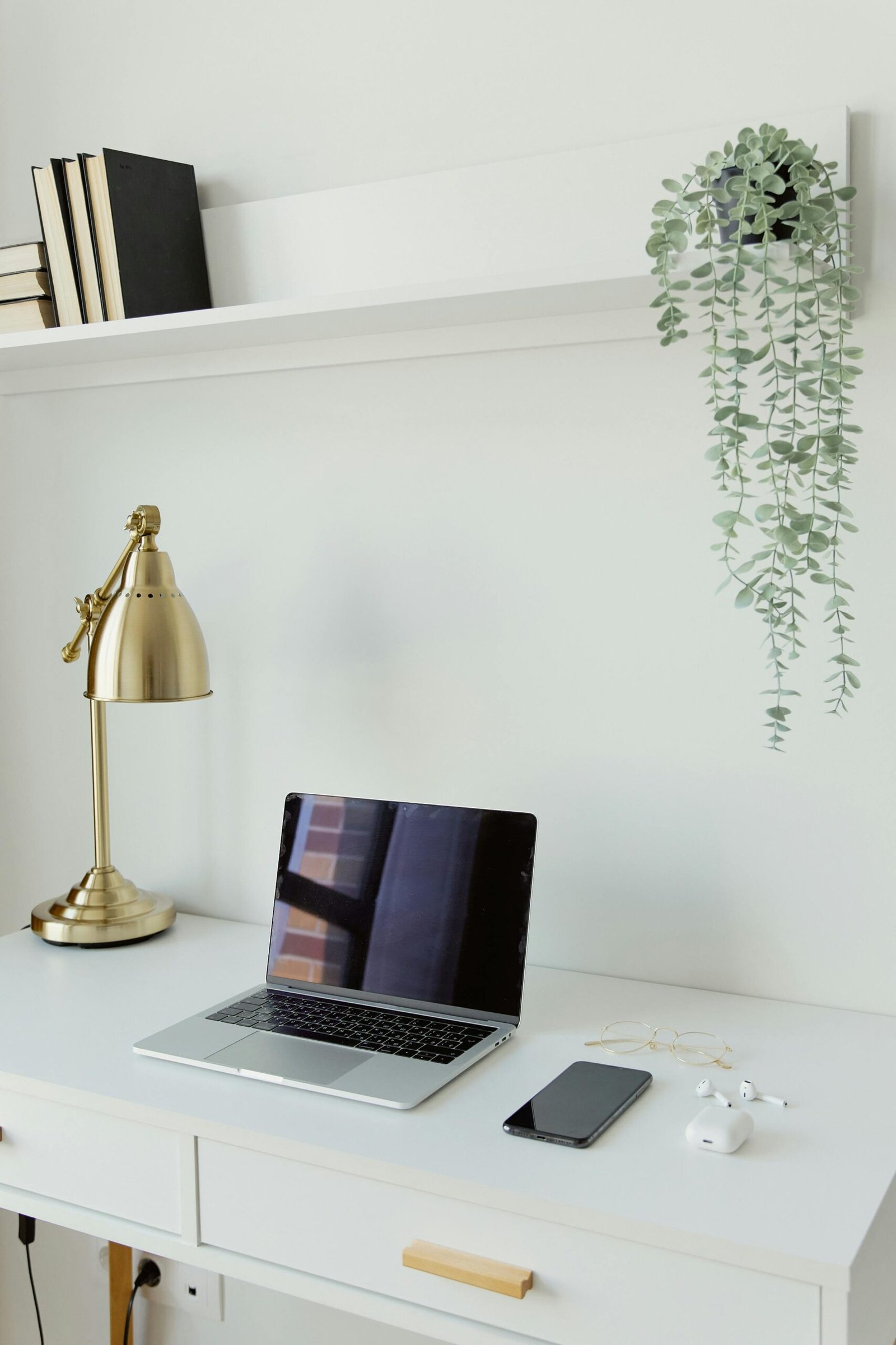 A modern, minimalist workspace featuring a laptop, smartphone, and brass lamp on a white desk.